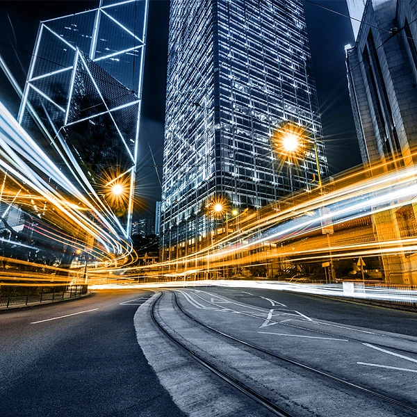 Eine nächtliche Stadtansicht zeigt eine belebte Straße mit verschwommenen Lichtern von Fahrzeugen, die eine dynamische Bewegungswirkung erzeugen. Im Hintergrund erhebt sich ein hochmodernes Gebäude mit einer hellen, reflektierenden Fassade.
