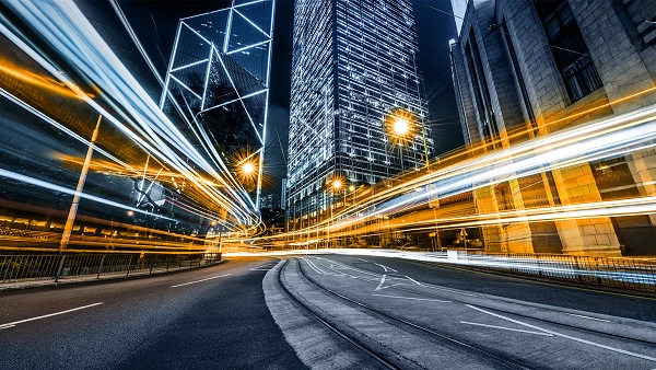 Eine nächtliche Stadtansicht zeigt eine belebte Straße mit verschwommenen Lichtern von Fahrzeugen, die eine dynamische Bewegungswirkung erzeugen. Im Hintergrund erhebt sich ein hochmodernes Gebäude mit einer hellen, reflektierenden Fassade.