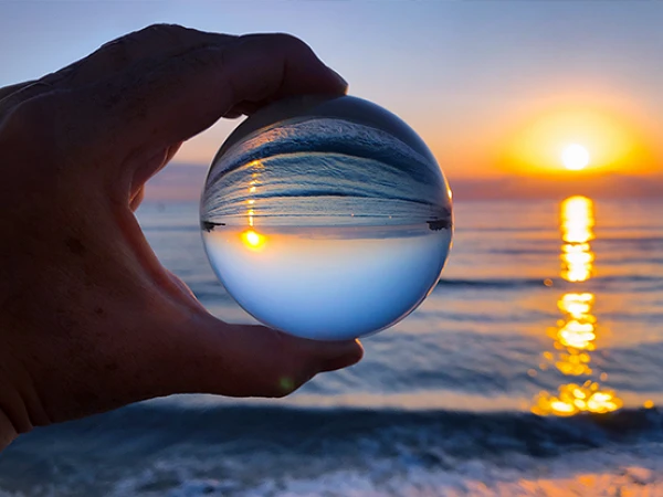 Eine Hand hält eine Glaskugel, in der der Sonnenuntergang über dem Meer reflektiert wird, während die Wellen sanft am Strand plätschern.