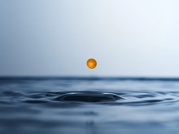 Eine orangefarbene Tropfen schwebt über der Wasseroberfläche und erzeugt Wellenripple, während der Hintergrund sanfte Blau- und Grautöne zeigt.