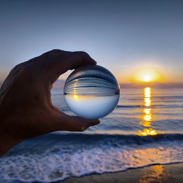 Eine Hand hält eine Glaskugel, die den Sonnenuntergang über dem Meer reflektiert, während sanfte Wellen an den Strand rollen.