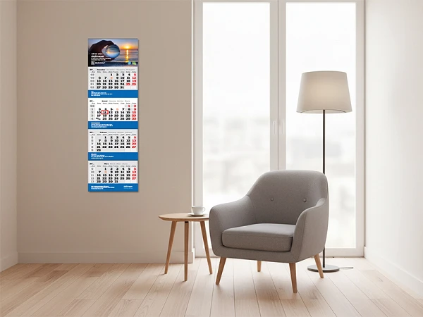 Ein Wandkalender mit mehreren Monaten ist an einer hellen Wand befestigt, während ein grauer Sessel und eine Stehlampe im Raum stehen. Ein Fenster lässt Tageslicht herein.