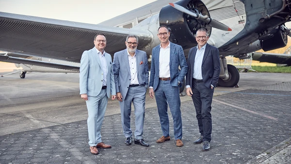 Vier Männer in eleganter Kleidung posieren lächelnd vor einem historischen Flugzeug auf einer gepflasterten Fläche. Das Flugzeug befindet sich im Hintergrund, und die Umgebung wirkt hell und freundlich.