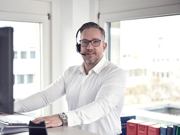Ein Mann in einem weißen Hemd sitzt an einem Schreibtisch in einem modernen Büro, trägt ein Headset und lächelt in die Kamera. Im Hintergrund sind Bücherregale mit verschiedenen Gesetzestexten zu sehen und ein Computerbildschirm befindet sich direkt vor ihm.
