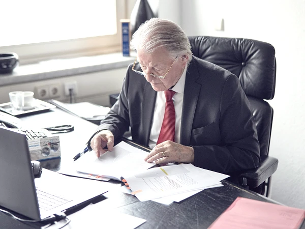 Ein älterer Mann mit grauem Haar und Brille sitzt an einem Schreibtisch und studiert Dokumente. Er trägt einen Anzug mit roter Krawatte und ist von Büromaterialien umgeben, während er auf einen Laptop schaut. Hinter ihm befindet sich ein helles Fenster.