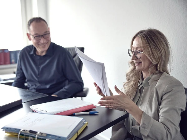 Eine lächelnde Frau mit Brille zeigt ein Dokument während eines Gesprächs mit einem Mann in einem modernen Büroraum, umgeben von Aktenordnern und einem Laptop.