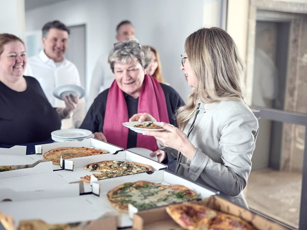 Eine Gruppe von Personen steht in einem Raum, während eine Frau mit blonden Haaren auf einen Teller mit Essen schaut, den sie in der Hand hält. Auf einem Tisch liegen mehrere Pizzakartons mit verschiedenen Pizzas. Im Hintergrund sind weitere Personen, die offensichtlich an der Essensausgabe teilnehmen. Die Atmosphäre wirkt freundlich und gesellig.