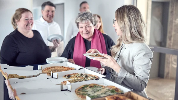 Eine Gruppe von Personen steht in einem Raum, während eine Frau mit blonden Haaren auf einen Teller mit Essen schaut, den sie in der Hand hält. Auf einem Tisch liegen mehrere Pizzakartons mit verschiedenen Pizzas. Im Hintergrund sind weitere Personen, die offensichtlich an der Essensausgabe teilnehmen. Die Atmosphäre wirkt freundlich und gesellig.