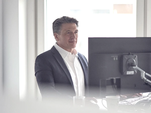 Ein Mann in einem dunklen Anzug steht an einem Schreibtisch mit einem Computerbildschirm, während er durch ein Fenster schaut. Im Hintergrund sind unscharfe Fenster und eine helle Umgebung zu sehen.