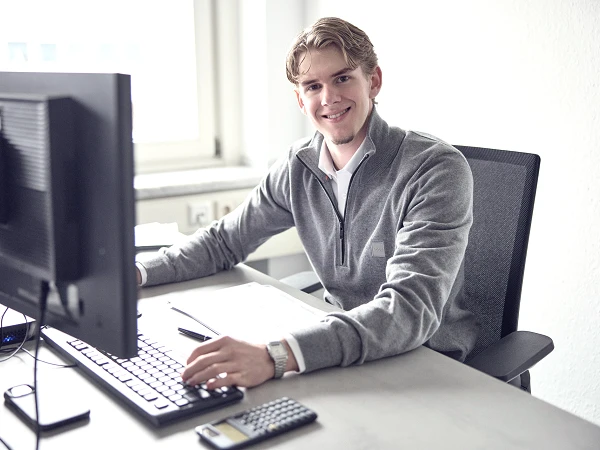 Ein junger Mann sitzt an einem Schreibtisch, lächelt in die Kamera und arbeitet an einem Computer. Auf dem Tisch liegen ein Notizblock und ein Taschenrechner.