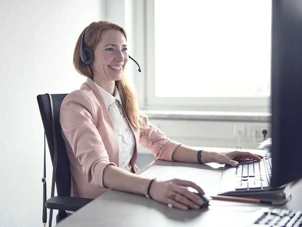 Eine lächelnde Frau mit Kopfhörern sitzt an einem Schreibtisch und arbeitet am Computer, während sie in einem hellen, freundlichen Raum sitzt.