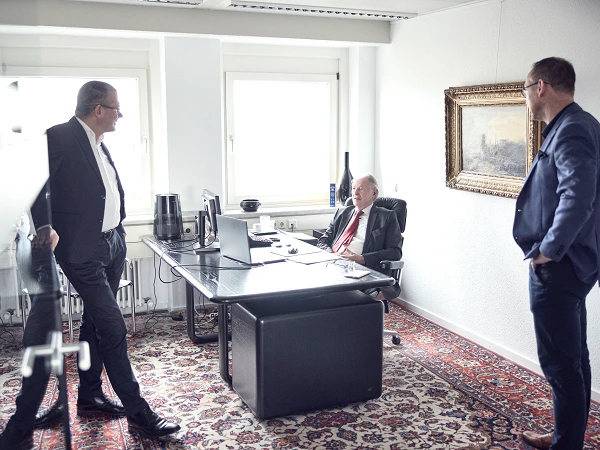 Drei Geschäftsmänner stehen in einem modernen Büroumfeld, zwei von ihnen sind im Gespräch, während der dritte, in einem Anzug mit Krawatte, hinter einem Schreibtisch sitzt und aufmerksam zuhört. An der Wand hängt ein Gemälde, und das Büro ist hell und minimalistisch eingerichtet.