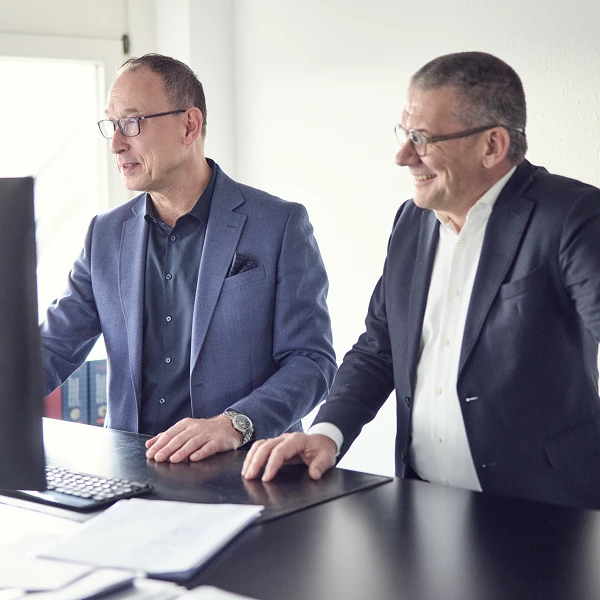 Zwei Männer in Anzügen stehen an einem Schreibtisch und schauen auf einen Computerbildschirm, während sie miteinander sprechen und lächeln.