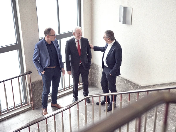 Drei Männer stehen auf einer Treppe und unterhalten sich freundlich, während sie an einem Geländer lehnen. Die Umgebung ist gut beleuchtet mit großen Fenstern, die Tageslicht hereinlassen.