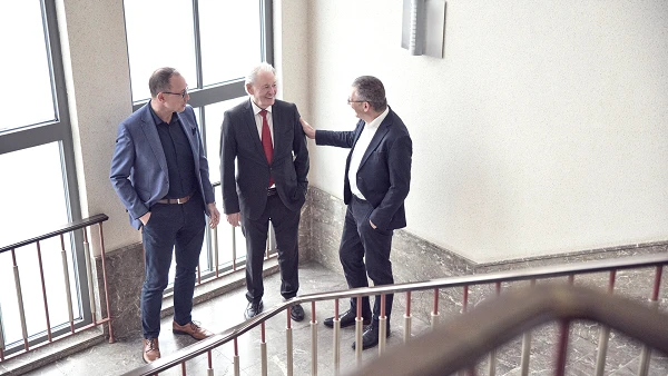 Drei Männer stehen auf einer Treppe und unterhalten sich freundlich, während sie an einem Geländer lehnen. Die Umgebung ist gut beleuchtet mit großen Fenstern, die Tageslicht hereinlassen.