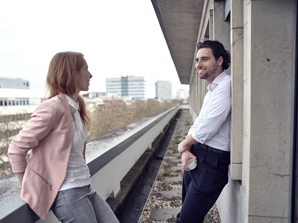Zwei Personen unterhalten sich entspannt auf einem Balkon, während sie urbanen Hintergrund mit modernen Gebäuden im Hintergrund genießen. Die Frau trägt eine hellrosa Blazerjacke und die Männer einen weißen Hemd, während er eine Tasse in der Hand hält.