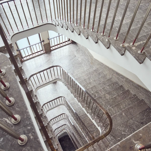 Eine moderne Treppe mit geschwungenen Geländern aus Metall, die in mehreren Ebenen nach unten führt. Die Stufen sind mit einem dunklen, marmorierten Bodenbelag versehen. Durch große Fenster fällt natürliches Licht in den Treppenraum.