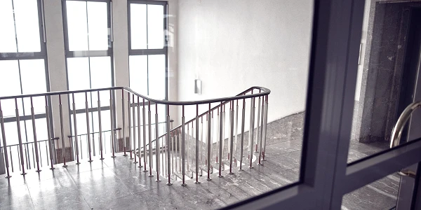 Eine schlichte Treppe mit einem gebogenen Geländer in einem hell erleuchteten Treppenhaus, umgeben von großen Fenstern. Der Boden ist mit grauem Marmor ausgelegt.