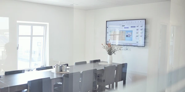 Ein heller, moderner Konferenzraum mit einem langen Tisch und Stühlen, ausgestattet mit einem Monitor an der Wand, auf dem eine Präsentation angezeigt wird. Ein Tisch mit Kaffeekannen und einer Blumenvase ist ebenfalls sichtbar.