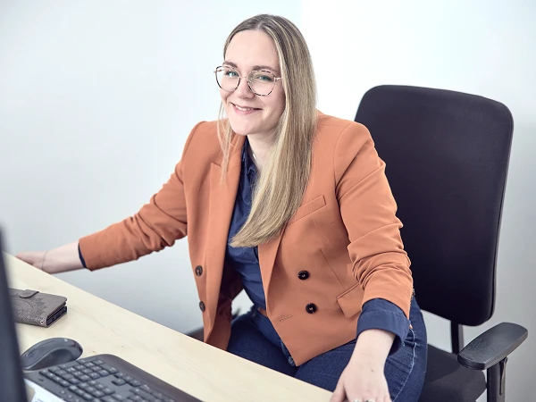 Eine Person mit langen blonden Haaren sitzt an einem Schreibtisch und lächelt in die Kamera. Sie trägt einen orangefarbenen Blazer über einem blauen Oberteil. Am Schreibtisch befinden sich ein Computer und eine Maus.