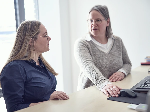 Zwei Frauen sitzen an einem Schreibtisch, wobei eine Frau mit langen, blonden Haaren in einem blauen Hemd die andere Frau mit grauen Haaren in einem hellen Pullover anblickt, während sie auf eine Maus und einen Computerbildschirm zeigen.