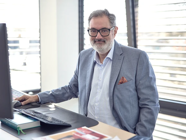 Ein Mann mit Brille und grauem Anzug sitzt an einem Schreibtisch mit Computer in einem modern gestalteten Büro, während er in die Kamera lächelt. Im Hintergrund sind Fenster mit horizontalen Jalousien sichtbar.