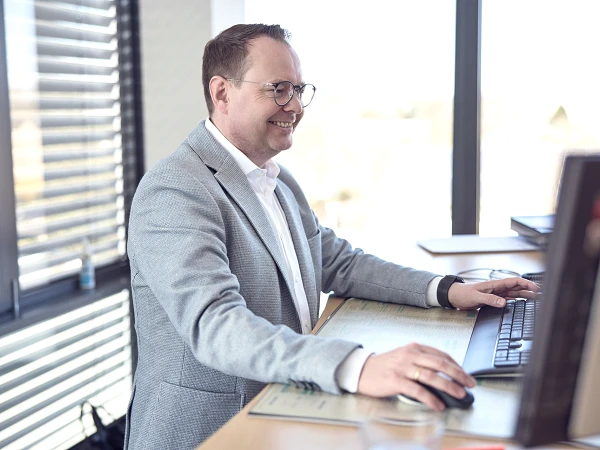 Ein Mann mit Brille sitzt an einem Schreibtisch, arbeitet an einem Computer und lächelt, während er auf die Tastatur schaut. Im Hintergrund sind Fenster mit Blick auf eine Stadtlandschaft.