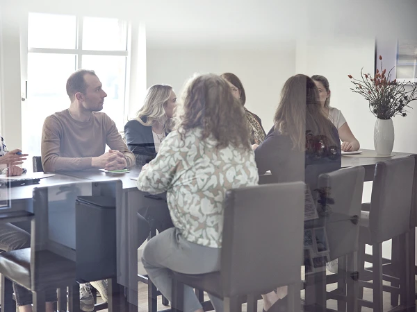 Eine Gruppe von fünf Personen sitzt an einem Tisch in einem hellen Raum, umgeben von Fenstern und einer Pflanze in einer Vase. Sie sind in ein Gespräch vertieft, während eine Person am Kopf des Tisches den anderen zuhört.