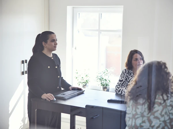 Eine Frau steht an einem Tisch mit einem Laptop, während zwei weitere Frauen am Tisch sitzen und diskutieren. Im Hintergrund sind Pflanzen und ein Fenster zu sehen, das Tageslicht hereinlässt.