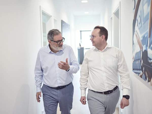 Zwei Männer in formeller Kleidung gehen durch einen hellen Büroflur und sprechen miteinander. Im Hintergrund sind Wandbilder und Bürotüren sichtbar.