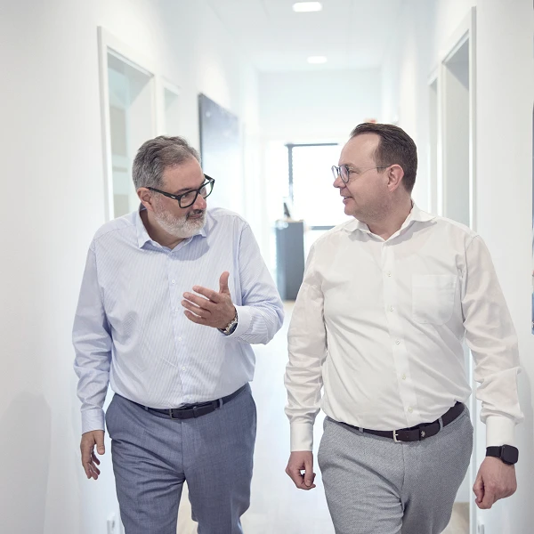 Zwei Männer in formeller Kleidung gehen durch einen hellen Büroflur und sprechen miteinander. Im Hintergrund sind Wandbilder und Bürotüren sichtbar.