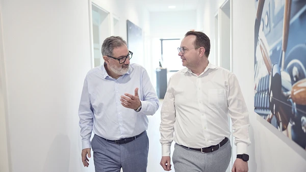 Zwei Männer in formeller Kleidung gehen durch einen hellen Büroflur und sprechen miteinander. Im Hintergrund sind Wandbilder und Bürotüren sichtbar.