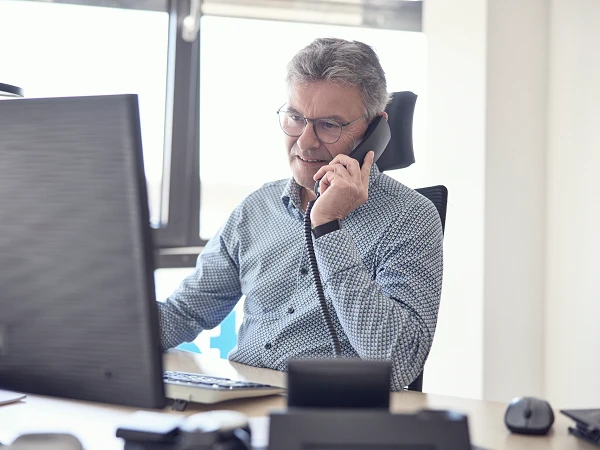 Ein Mann mit Brille und kurzem grauem Haar führt ein Telefongespräch, während er an einem Schreibtisch sitzt und auf einen Computerbildschirm schaut.