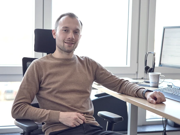 Ein Mann sitzt entspannt in einem Bürostuhl vor einem Schreibtisch mit einem Computerbildschirm, er trägt einen braunen Pullover und lächelt in die Kamera. Im Hintergrund sind Fenster mit Blick auf eine leicht verschwommene Außenansicht zu sehen.