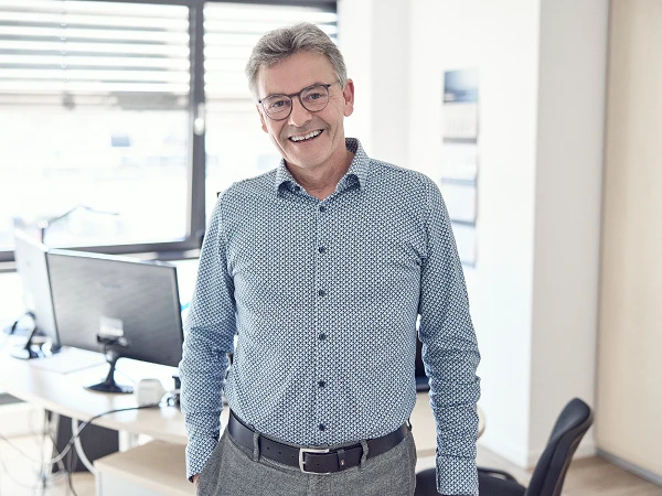 Ein lächelnder Mann mit Brille steht in einem hellen Büro, hinter ihm sind Computer und Büromöbel zu sehen. Er trägt ein blaues, gemustertes Hemd und graue Hosen.