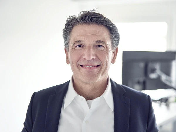 Ein lächelnder Mann mit grauen Haaren trägt einen dunkelblauen Blazer und ein weißes Hemd. Er steht in einem Büro, im Hintergrund ist ein Bildschirm sichtbar.