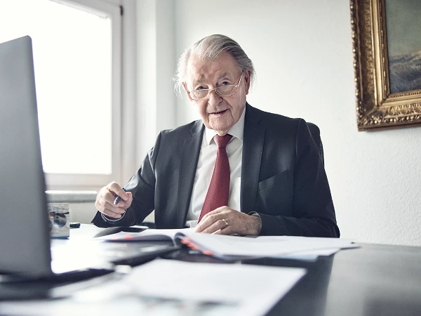 Ein älterer Mann mit Brille und grauen Haaren sitzt an einem Schreibtisch, trägt einen schwarzen Anzug und eine rote Krawatte. Er schaut direkt in die Kamera und hat einen Stift in der Hand, während Dokumente vor ihm auf dem Tisch liegen. Im Hintergrund ist ein Fenster und ein eingerahmtes Bild zu sehen.