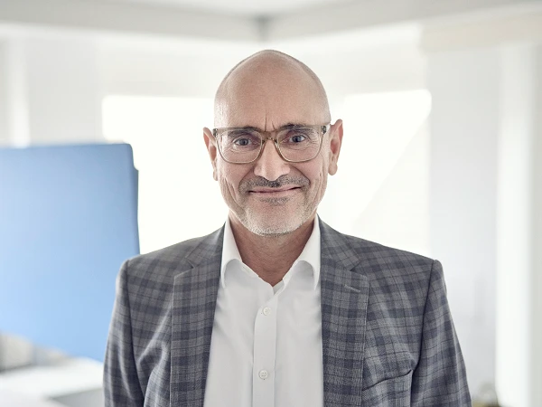 Ein lächelnder Mann mittleren Alters trägt eine Brille und einen karierten Anzug und steht in einem hellen, modernen Raum mit einem blauen Hintergrund.