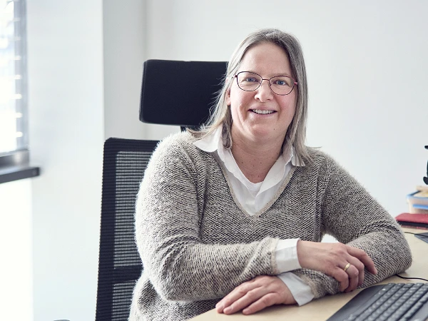 Eine lächelnde Frau mit Brille sitzt an einem Schreibtisch, auf dem ein Computer und einige Bücher liegen. Sie trägt einen beigen Pullover über einem weißen Hemd und hat ihr Haar offen.