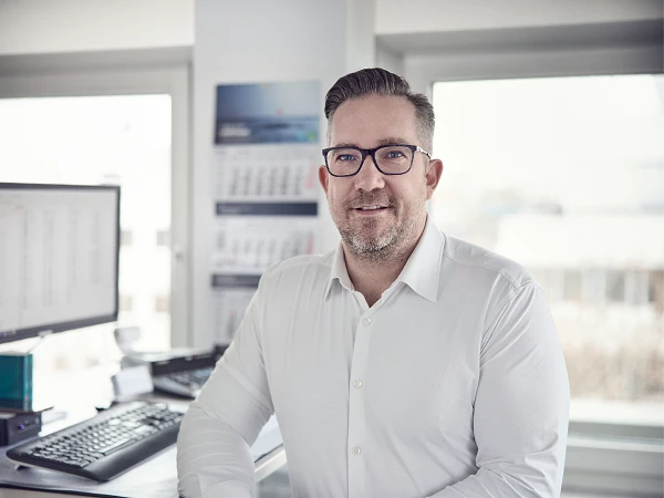 Ein Mann mit Brille und einem weißen Hemd steht in einem Büro, während er an einem Schreibtisch mit einem Computer und einer Tastatur lehnt, im Hintergrund sind Fenster mit Tageslicht und eine Wand mit einem Kalender zu sehen.