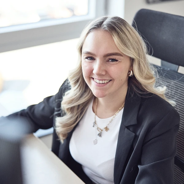 Eine junge Frau mit blonden, lockigen Haaren sitzt an einem Schreibtisch und lächelt freundlich in die Kamera. Sie trägt ein schwarzes Blazer und einen weißen Shirt, während sie vor einem Computerbildschirm sitzt. Im Hintergrund sind Fenster zu sehen, die Tageslicht hereinlassen.