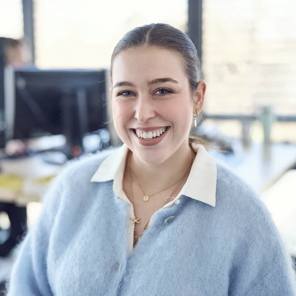 Eine lächelnde junge Frau mit einem hellblauen Pullover und einer weißen Bluse steht in einem modernen Büro vor einem Computerarbeitsplatz.