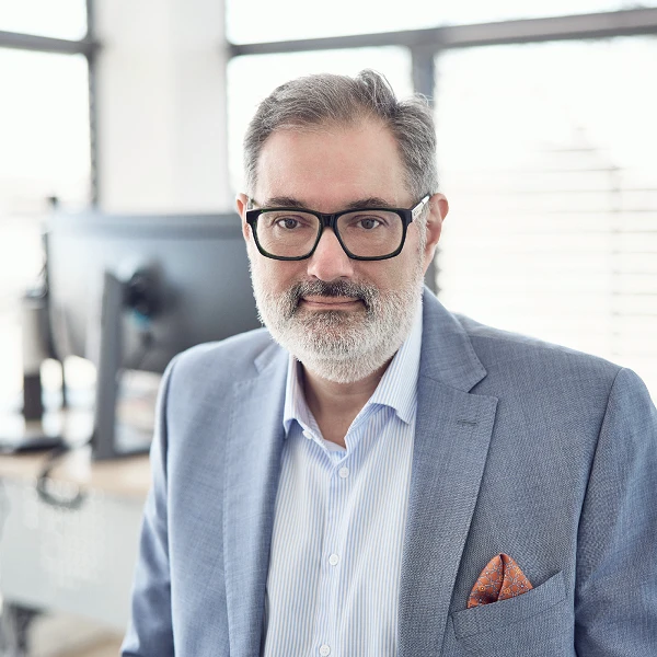 Ein Mann mittleren Alters mit grauem Bart und Brille, trägt einen hellblauen Anzug und ein gestreiftes Hemd, steht in einem modernen Büro mit großen Fenstern im Hintergrund.