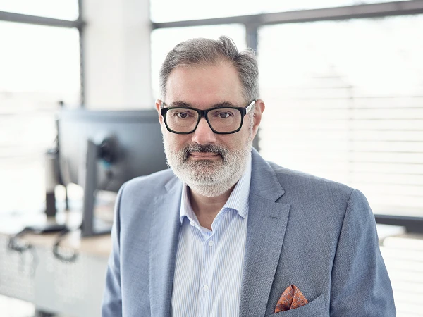 Ein Mann mittleren Alters mit grauem Bart und Brille, trägt einen hellblauen Anzug und ein gestreiftes Hemd, steht in einem modernen Büro mit großen Fenstern im Hintergrund.