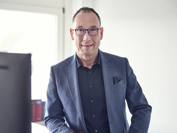 Ein lächelnder Mann in einem eleganten blauen Anzug steht an einem Schreibtisch und schaut in die Kamera, während er von einem Fenster mit natürlichem Licht beleuchtet wird.