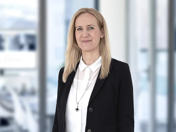 Eine blondhaarige Frau mit mittellanger Haarlänge trägt eine schwarze Jacke über einer weißen Bluse und steht in einem modernen, hellen Büro mit großem Fenster im Hintergrund.
