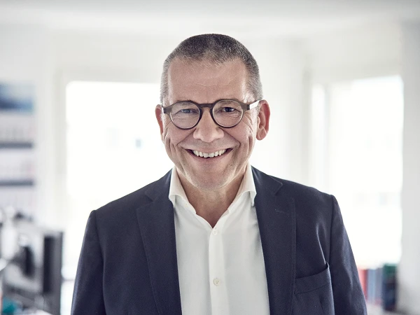 Ein lächelnder Mann mit Brille trägt einen dunklen Anzug und ein weißes Hemd, steht in einem hellen Raum mit Büromöbeln im Hintergrund.