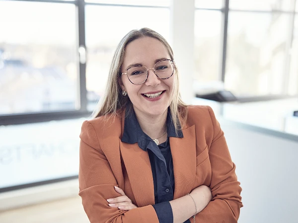 Eine Frau steht mit verschränkten Armen vor großen Fenstern und lächelt. Sie trägt ein orangefarbenes Blazer-Jacket und eine blaue Bluse. Im Hintergrund sind verschwommene Büroelemente zu erkennen.