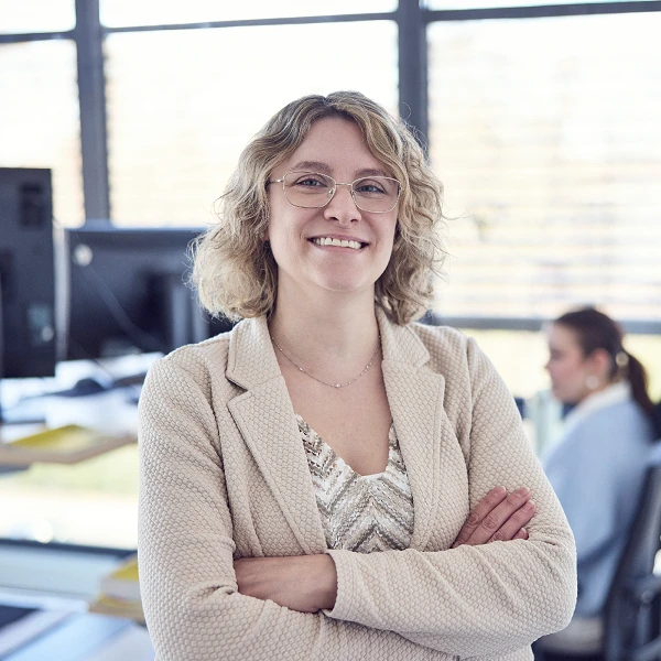 Eine lächelnde Frau mit lockigem, blondem Haar und Brille steht selbstbewusst mit verschränkten Armen in einem modernen Büro, im Hintergrund sind zwei weitere Mitarbeiterinnen zu sehen.
