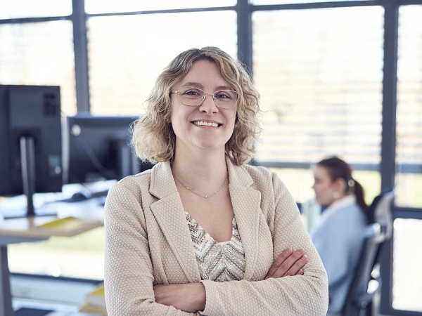 Eine lächelnde Frau mit lockigem, blondem Haar und Brille steht selbstbewusst mit verschränkten Armen in einem modernen Büro, im Hintergrund sind zwei weitere Mitarbeiterinnen zu sehen.